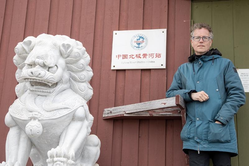 Ein mann henslengt foran ein raud bygning. Ved sidan han ein dør, og ved sisan av den igjen ein statue av ein løve i kinesisk stil.